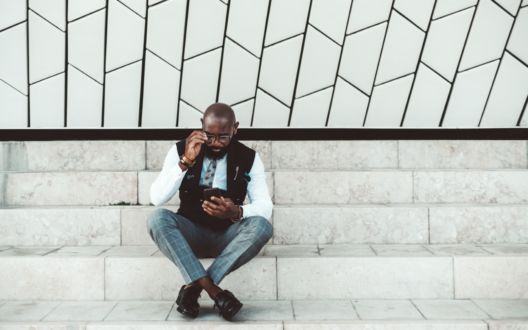 A fancy bearded bold African man entrepreneur is fixing his glasses and looking at the screen of his smartphone while sitting on the marble steps, with a copy space place on the right for your ad text